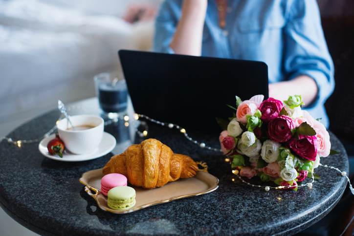 Young woman freelancer in blue shirt working on laptop during lunch with coffee, croissant, macaroons and flowers on table
